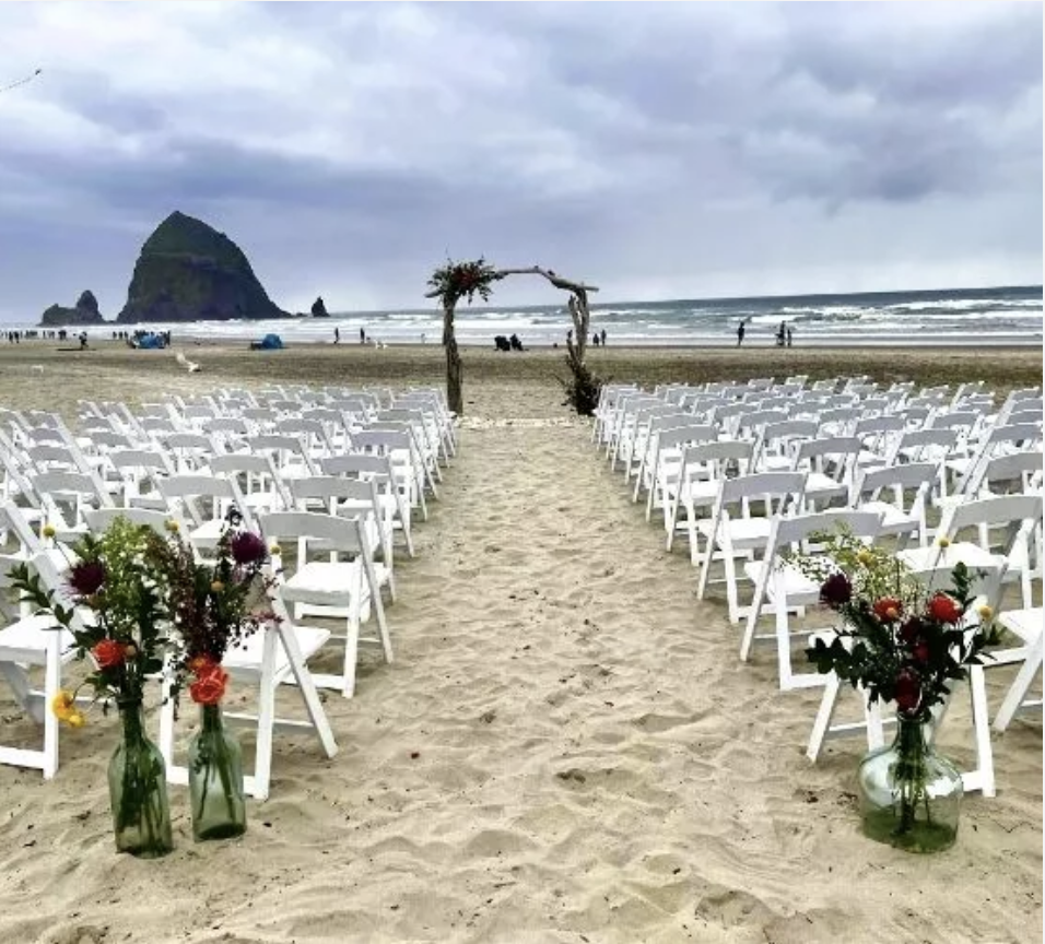White ceremony chairs arranged on a sandy beach with Haystack Rock in the background under a cloudy sky.